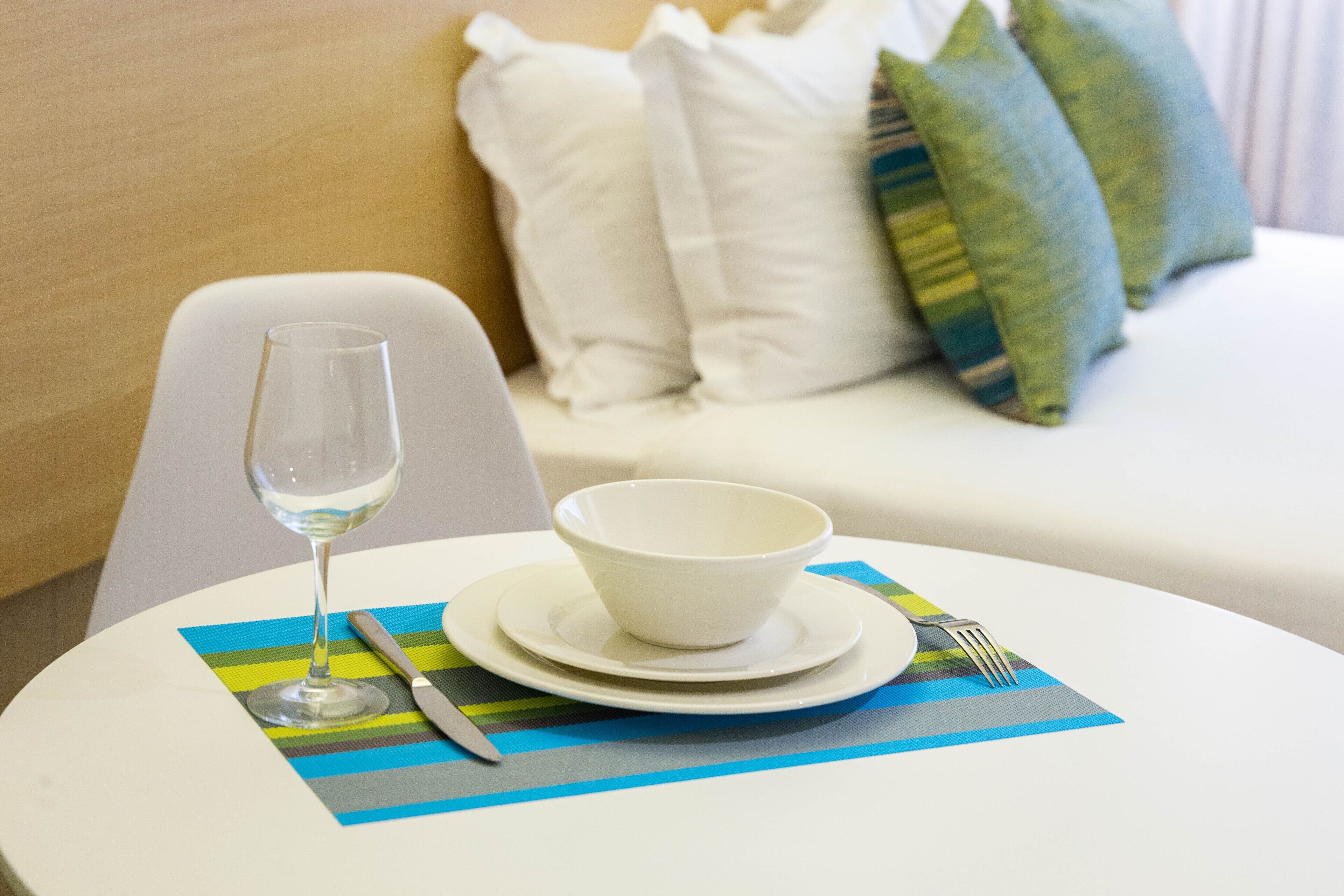 A dining setup with a white table, a bowl, a wine glass, and cutlery on a striped placemat.