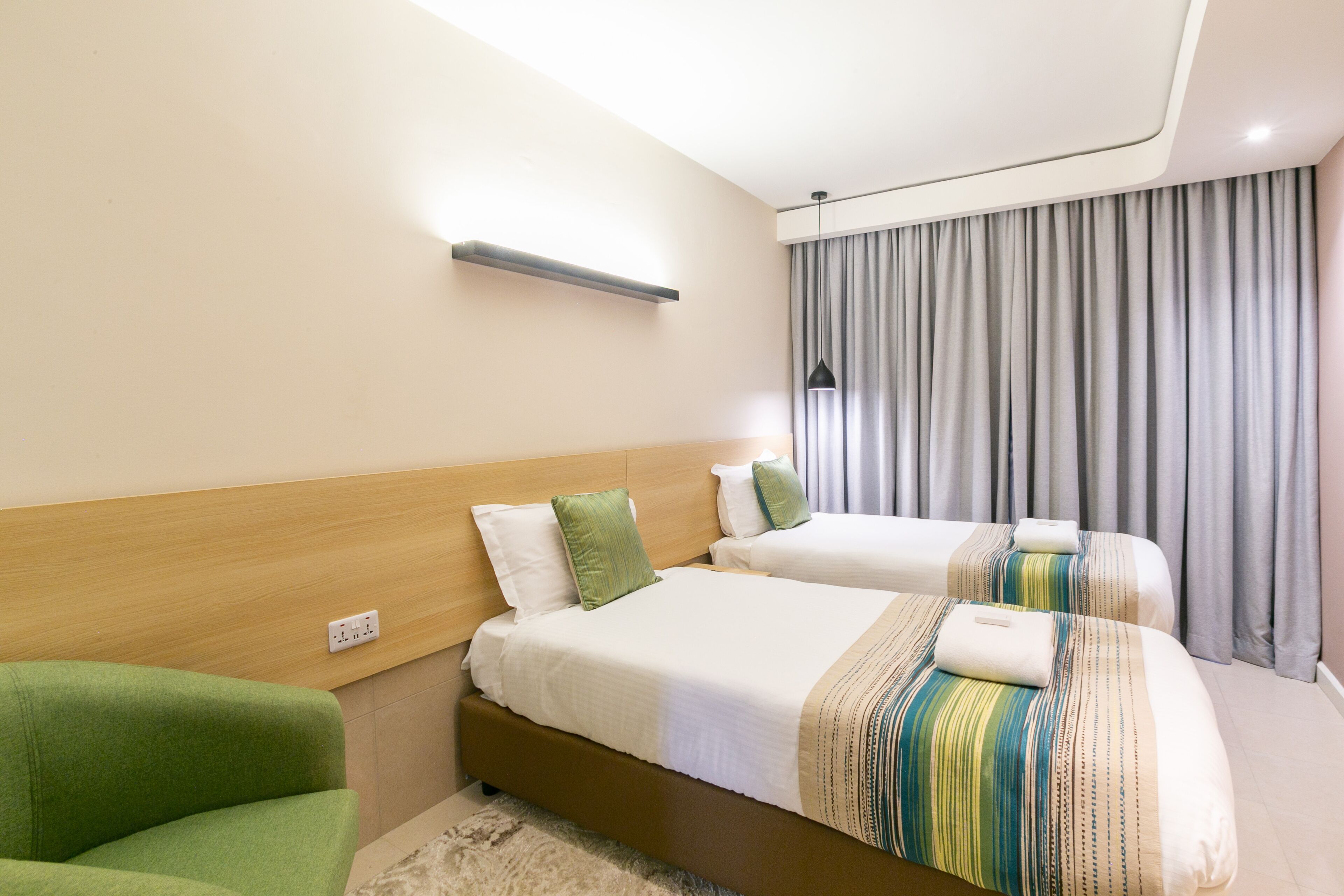 Habitación de hotel con dos camas, un sillón verde, cabecera de madera, un interruptor de luz y una ventana con cortinas grises.