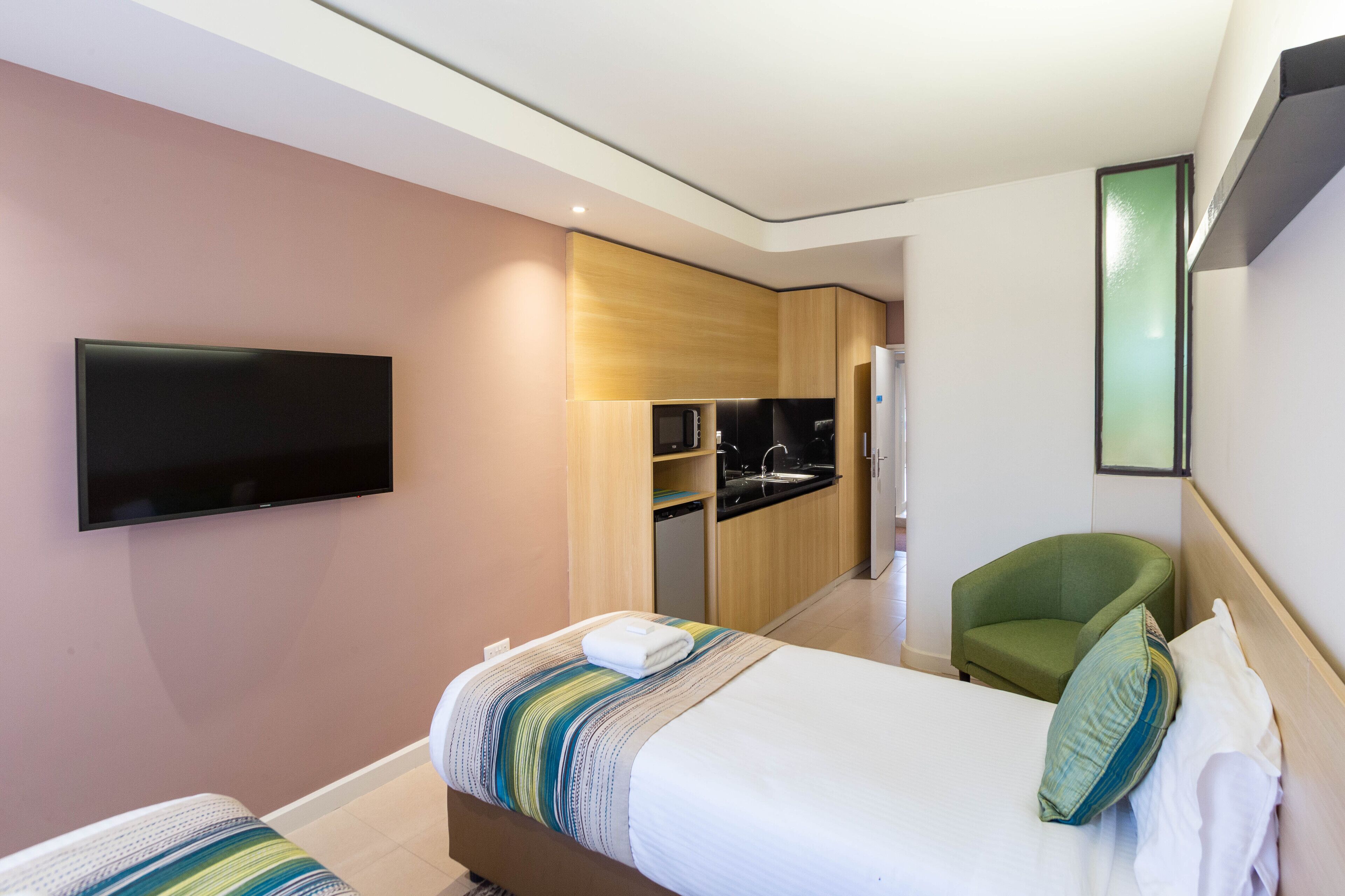 A modern hotel room with a bed, a green armchair, a flat-screen TV, and a kitchenette.