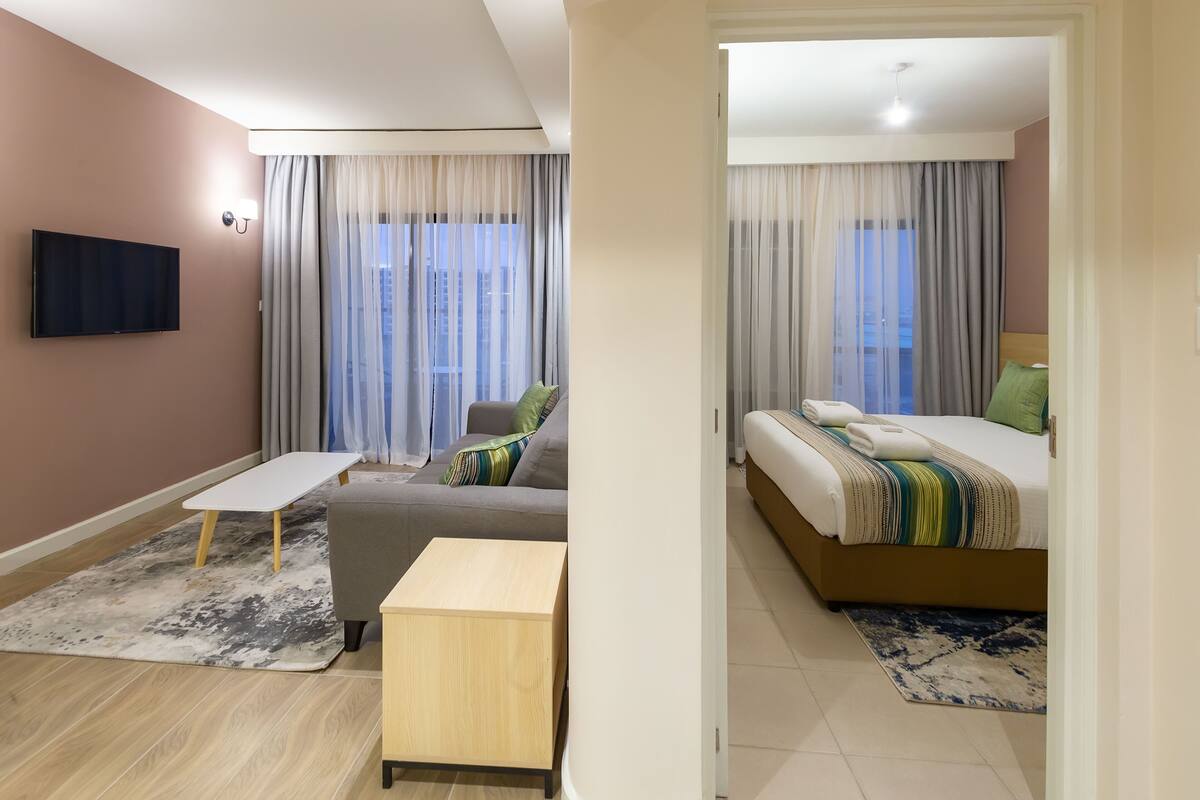 A modern hotel room with a sofa, a small wooden table, a bed with a striped blanket, and a TV mounted on the wall.
