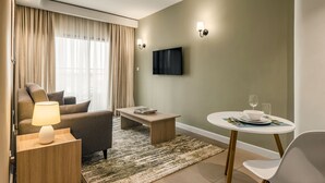 A modern hotel room with a sofa, a small table with a lamp, a TV mounted on the wall, and a dining area with a table set for two.