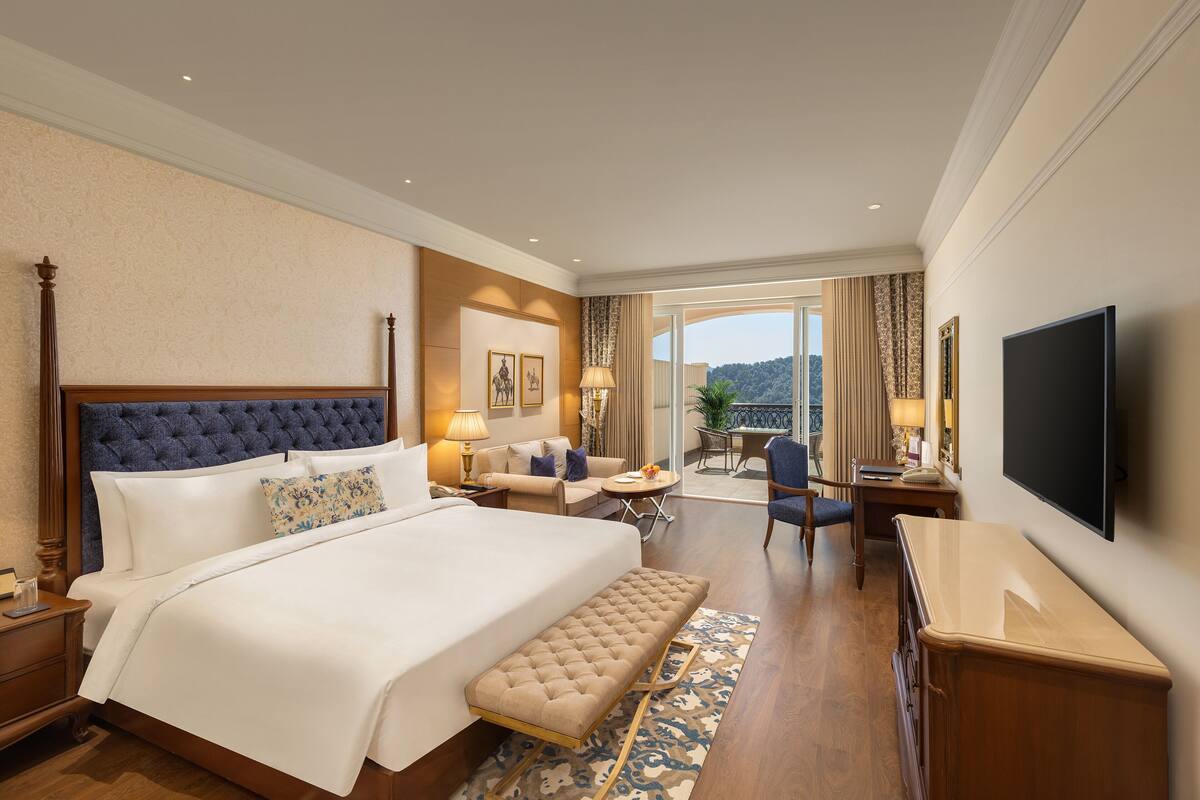 A hotel room with a large bed, a sitting area with a sofa and armchairs, a television, and a balcony with a view of the hills.