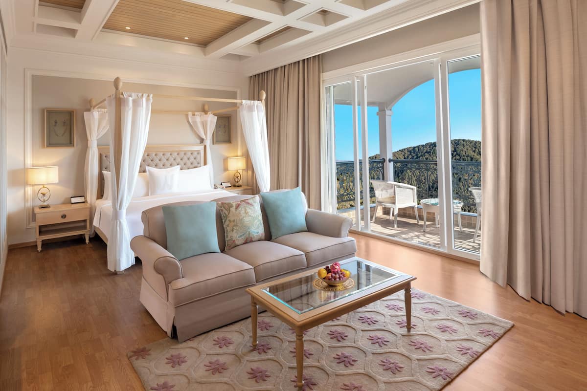A bedroom with a four-poster bed, a sofa, a glass coffee table, and a balcony with outdoor furniture.