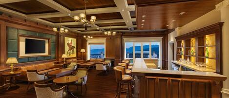 A spacious bar area with a high ceiling and wooden beams, a seating area with leather chairs and tables, and a large window offering a view of the outdoors.