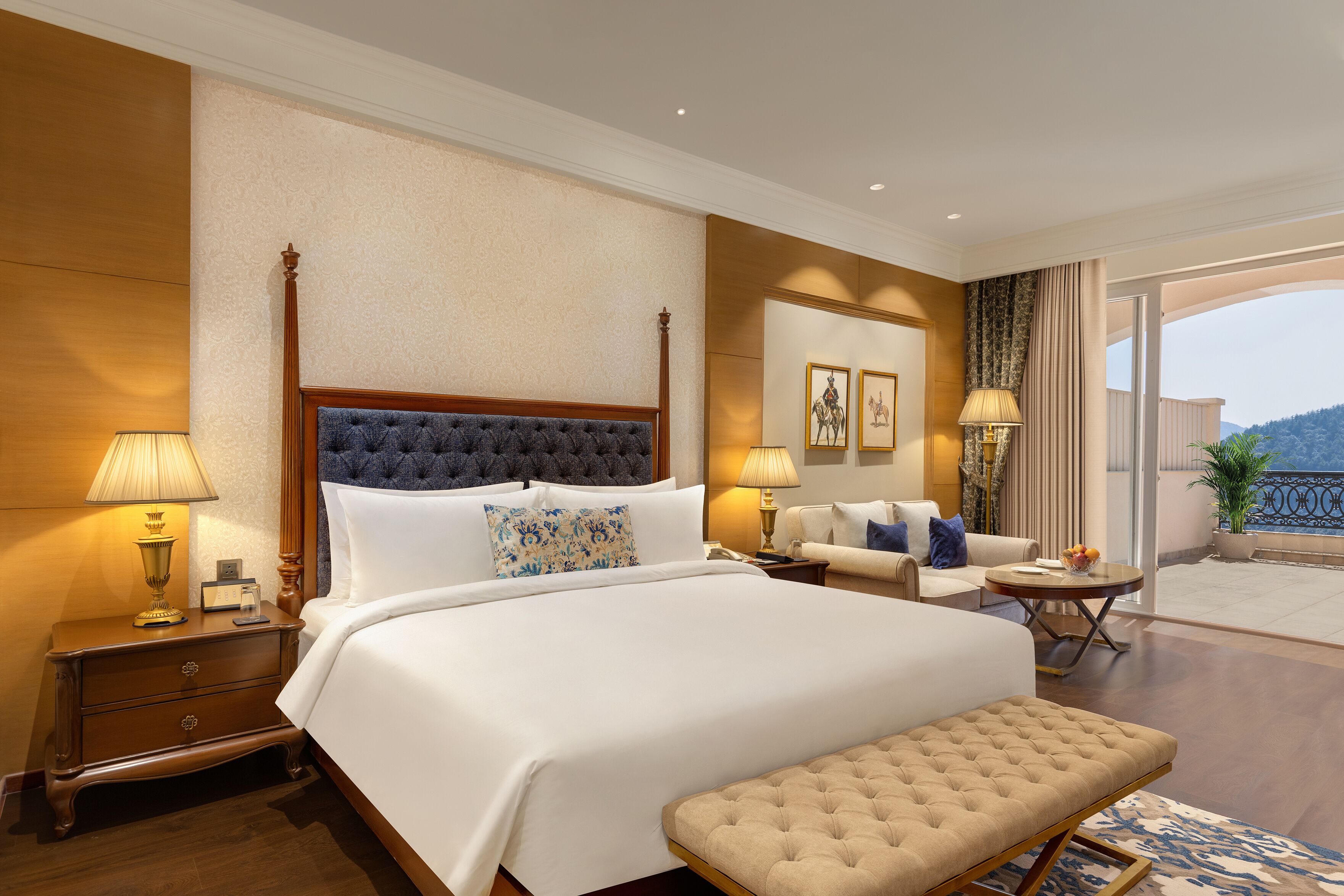 A hotel room with a large bed, bedside tables, a sofa, a small table, and a balcony with a view.