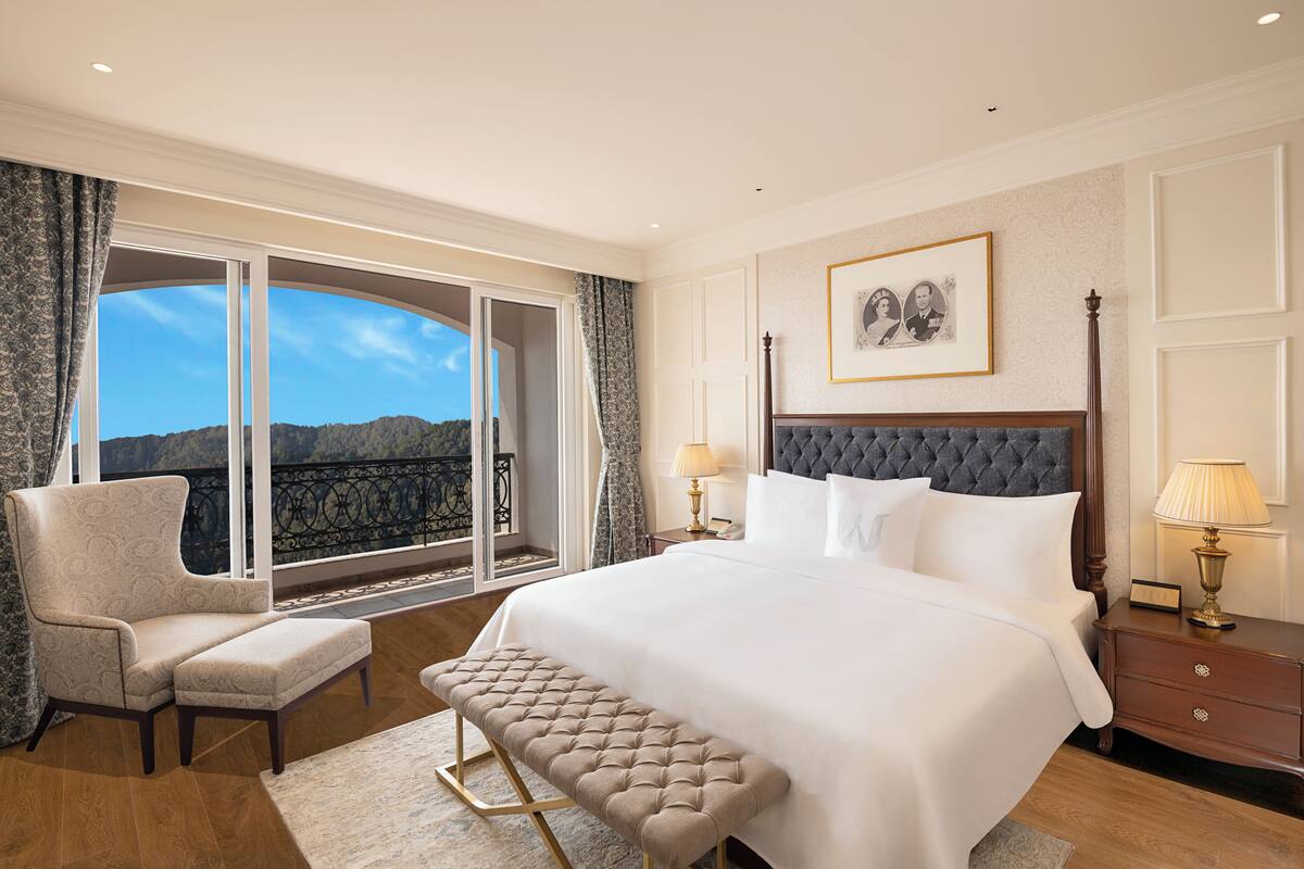 A spacious bedroom with a large bed, a bench, a chair, and a view of the mountains through a large window.