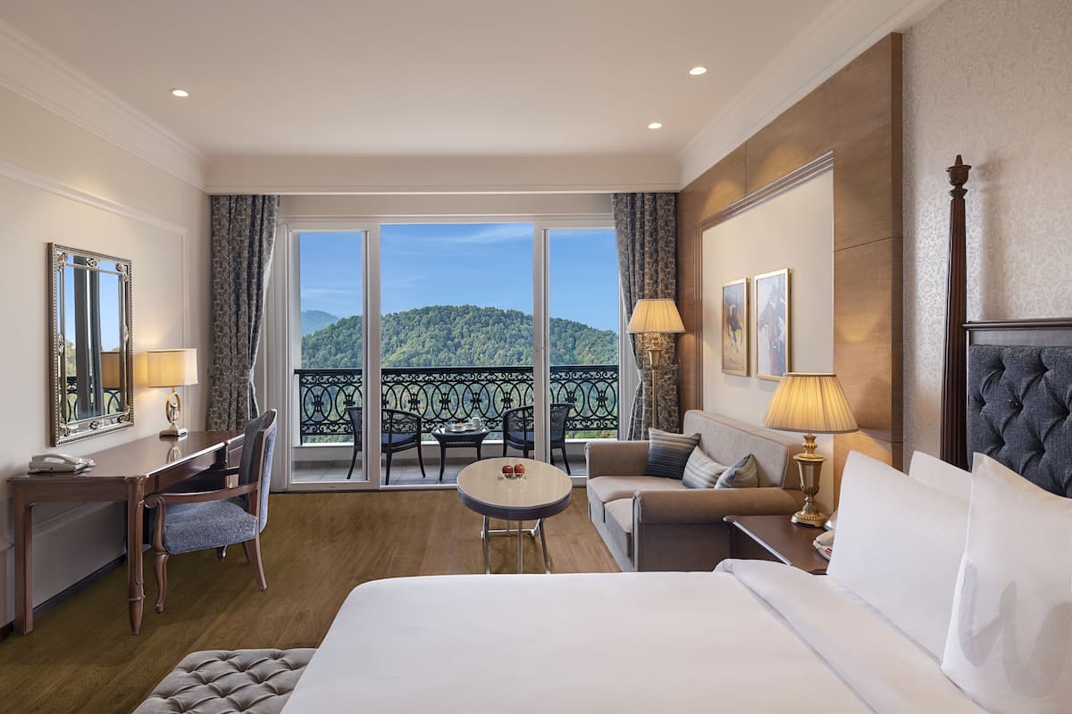 A hotel room with a large bed, a desk, a chair, a small round table, and a balcony with a view of a green hill.