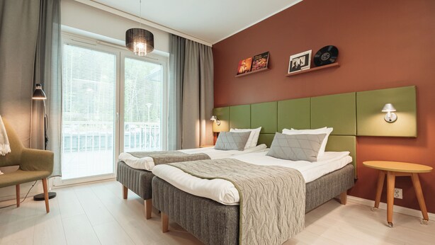 A hotel room with a large bed, a chair, a small table, and a window with blinds. - Time Apartments Ylistönmäki (Jyväskylä)