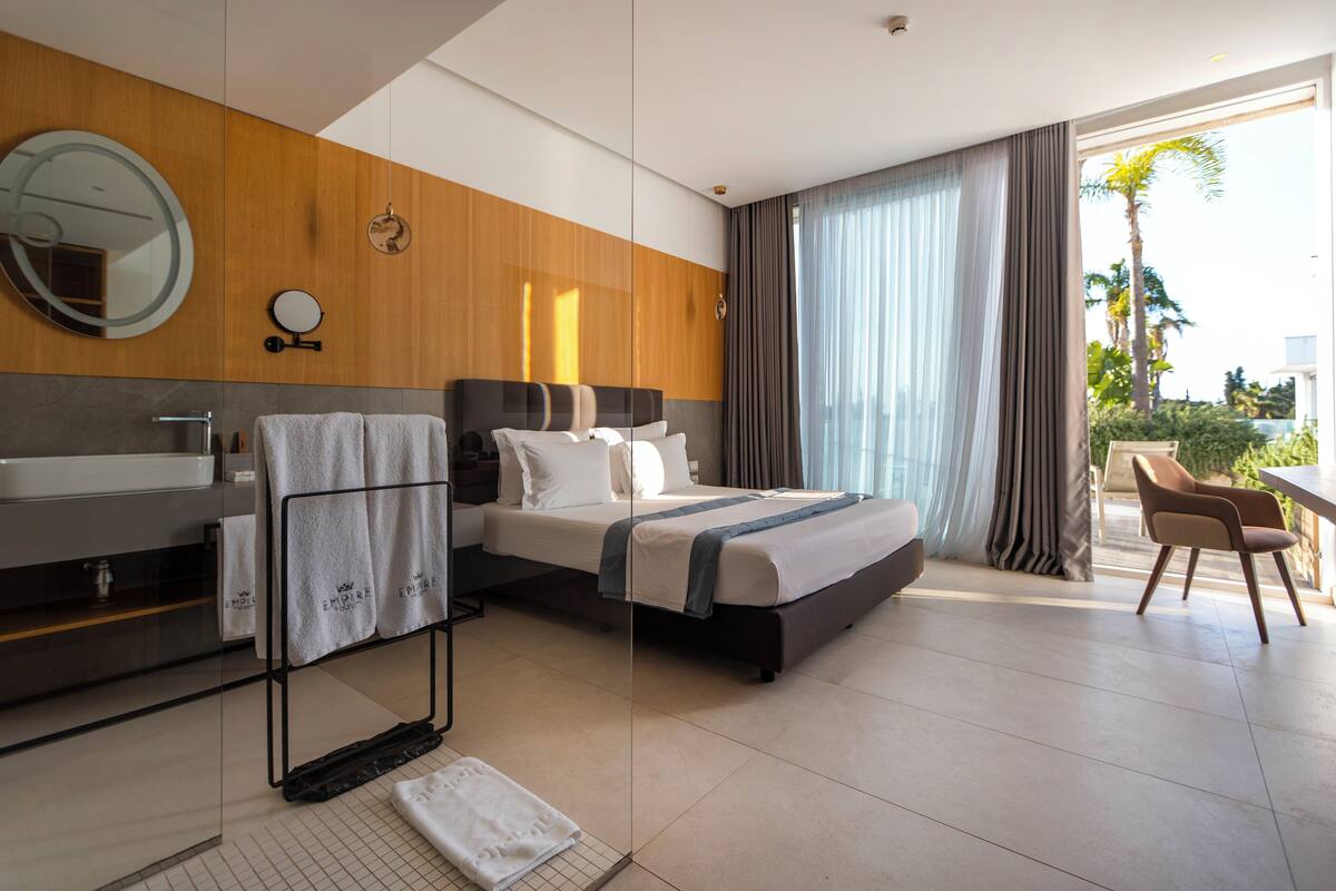 Una habitación de hotel moderna con una cama grande, un baño con lavamanos y espejo, y vistas a palmeras.