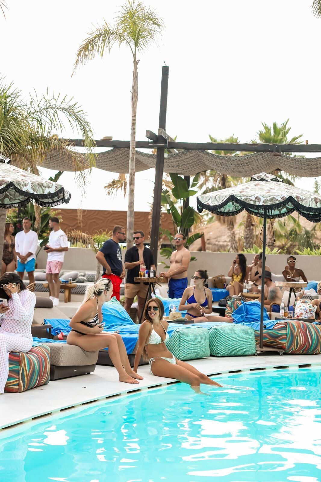 A poolside area with lounge chairs and umbrellas, surrounded by palm trees and a clear pool.