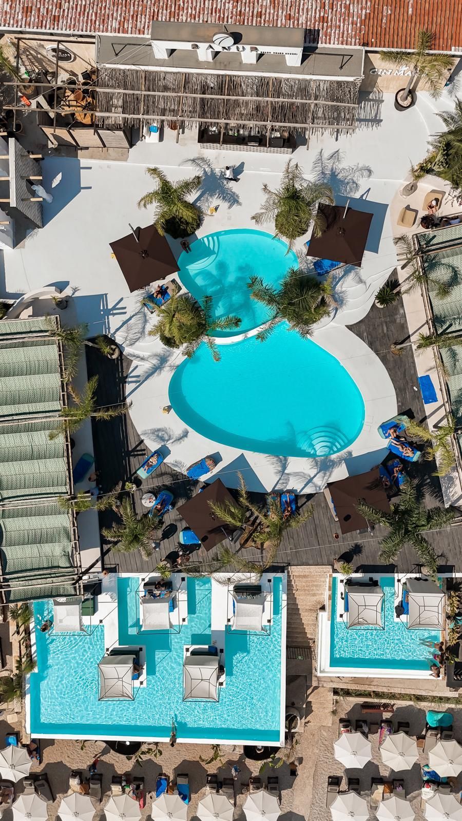 Vue aérienne d’un complexe hôtelier doté d’une piscine centrale entourée de chaises longues et de parasols, ainsi que de plusieurs petites piscines et de palmiers.