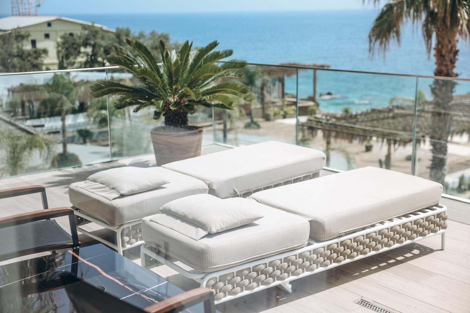 A balcony with three white cushioned lounge chairs, a glass table, and a potted plant.