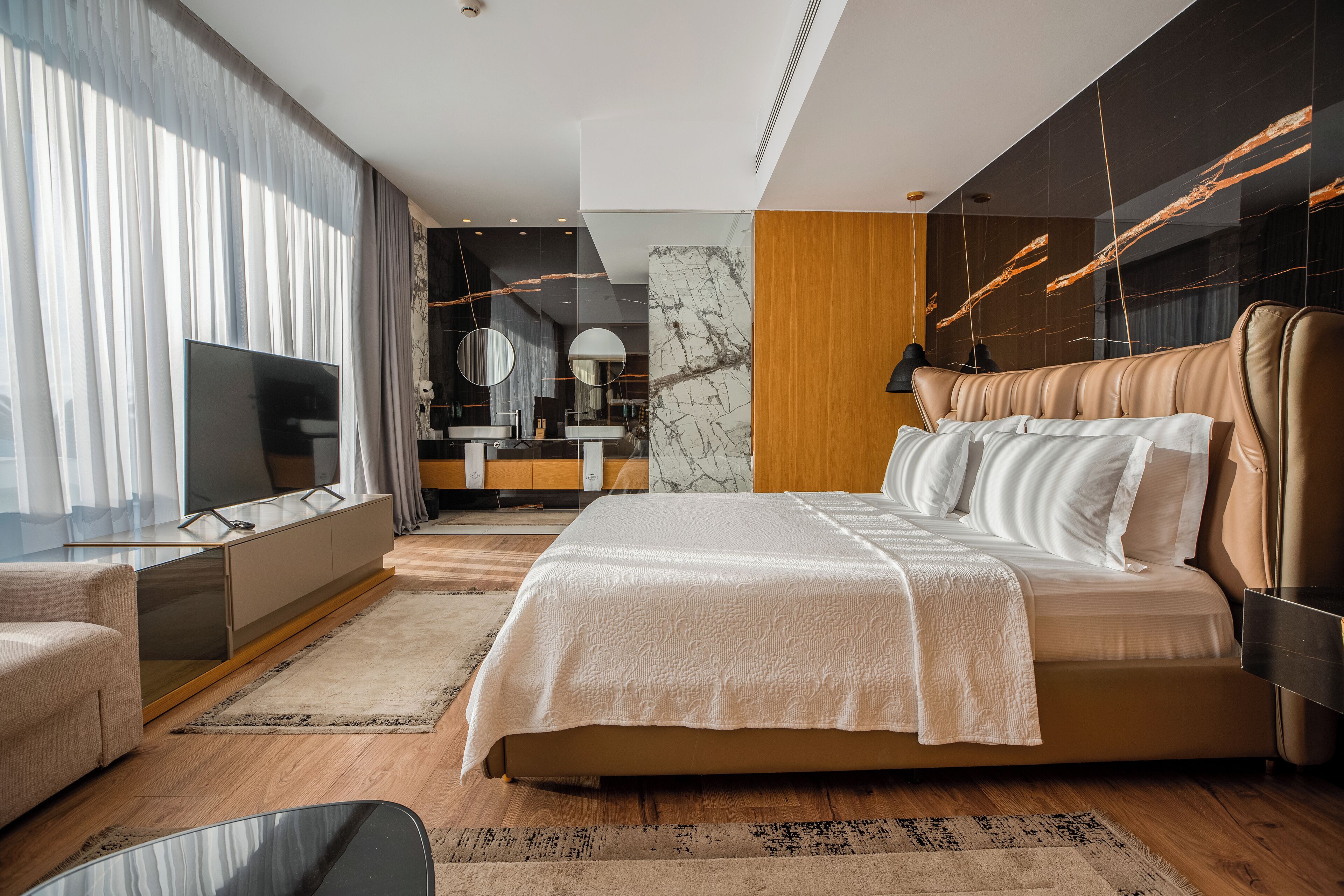 A modern bedroom with a large bed, a TV, a vanity, and a bathroom in the background.