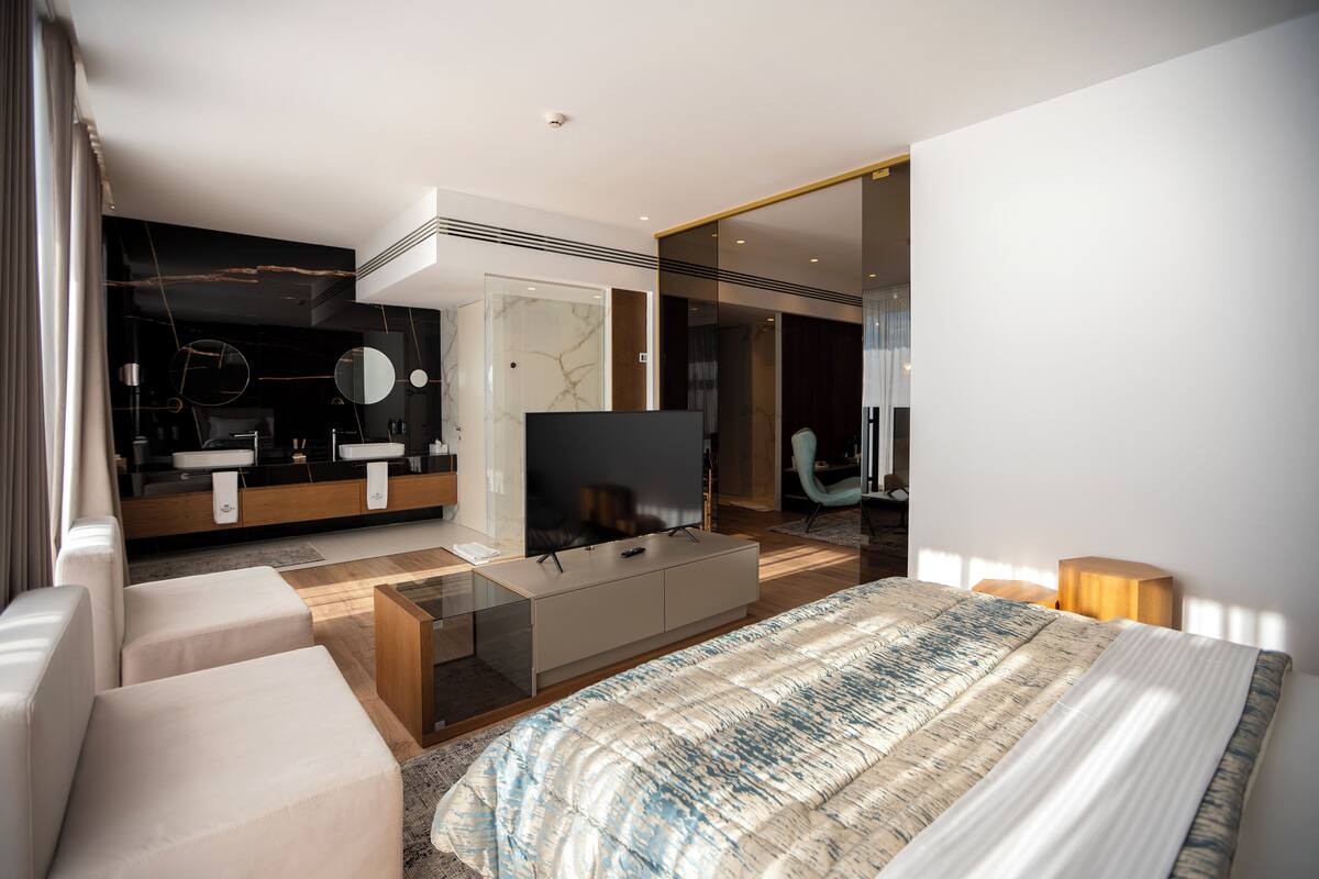 Una habitación de hotel moderna con una cama grande, televisor, sofá y baño con espejo y lavamanos.