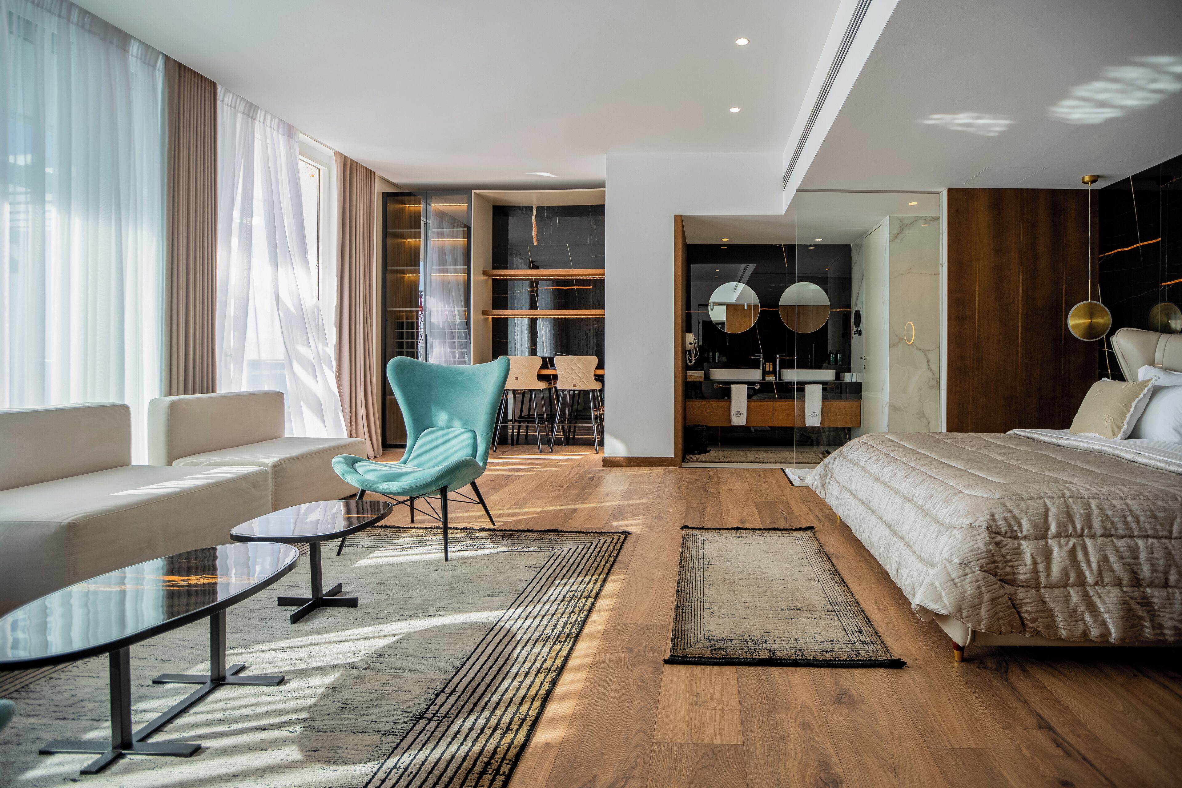A modern hotel room with a sofa, a teal chair, a bed, a small table, and a bathroom visible through an open door.