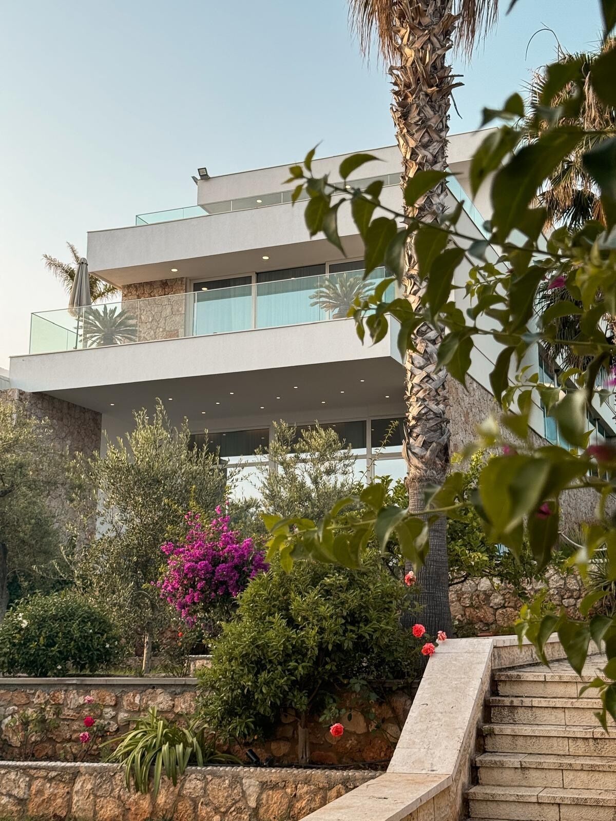 A modern multi-story building with balconies, surrounded by lush greenery and flowering plants.