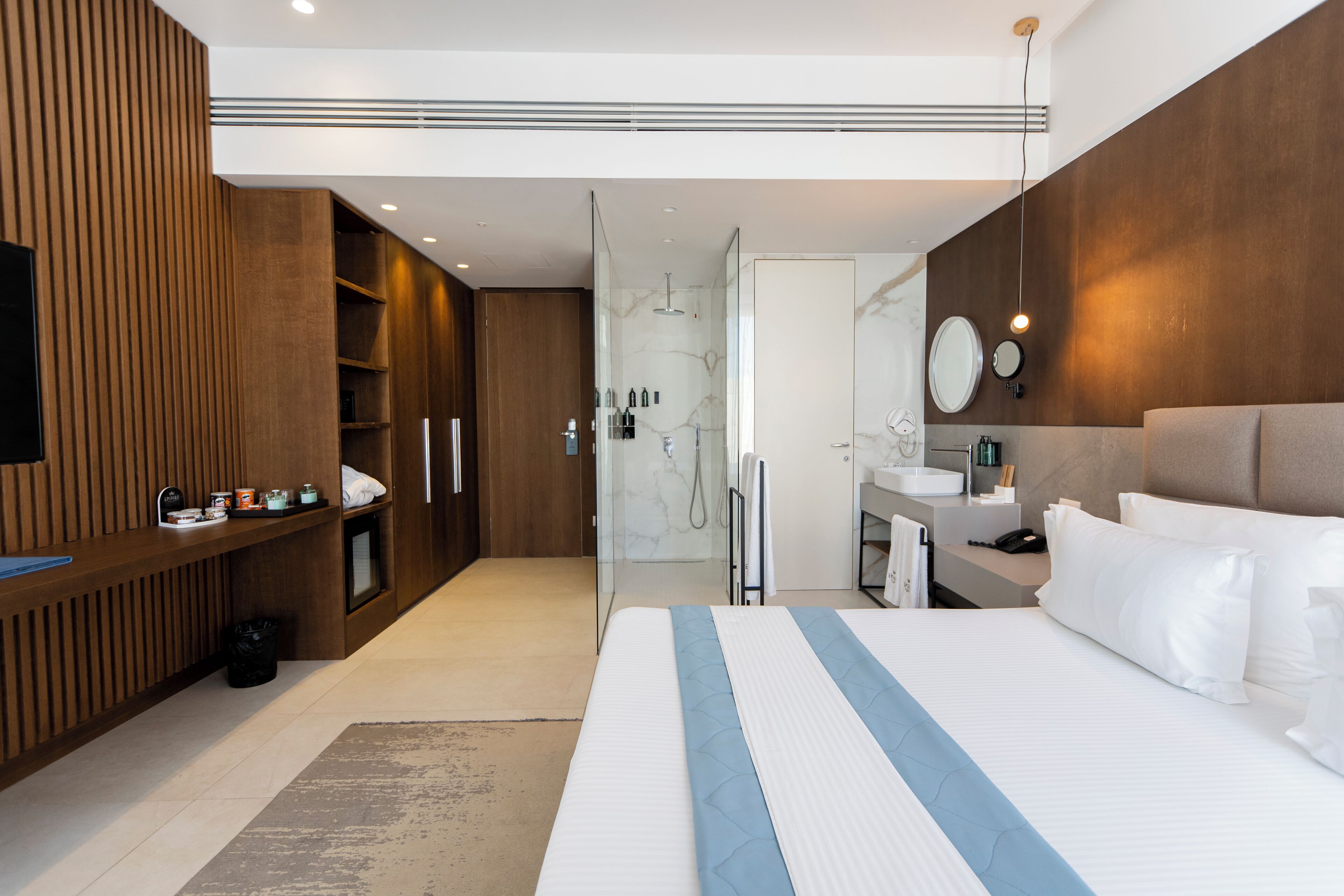 A modern hotel room with a large bed, a vanity with a mirror, a shower area, and a wooden panel wall.