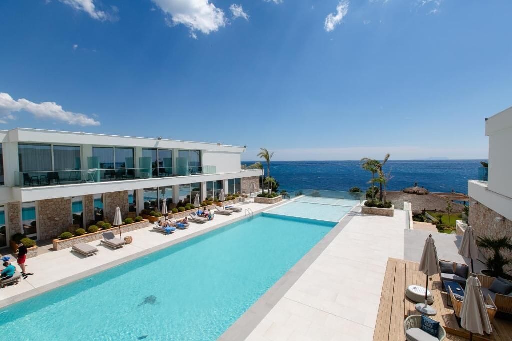 A modern hotel with a large infinity pool overlooking the ocean, lounge areas, and a clear view of the sea.