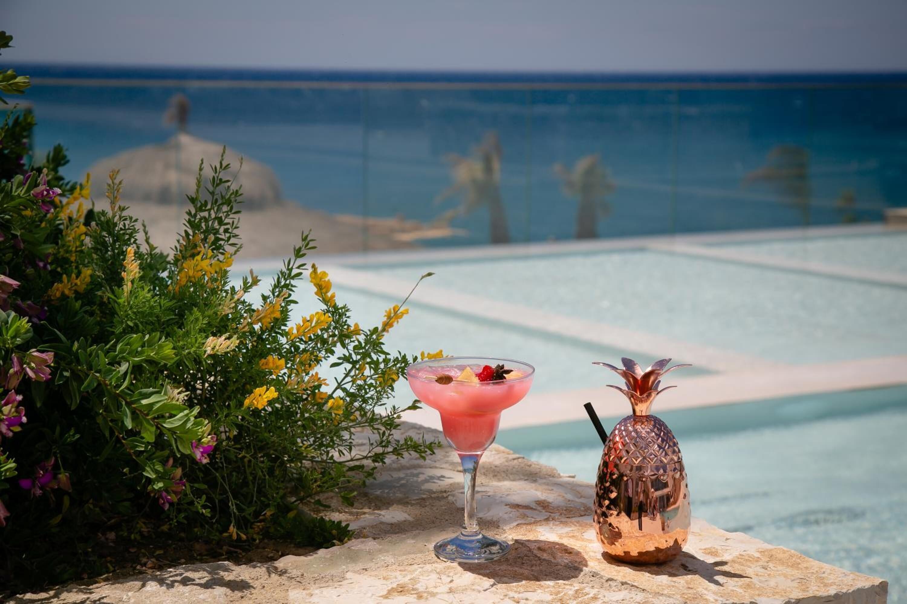 Un cóctel sobre un borde de piedra con una decoración de piña y una vista despejada de una piscina y el océano al fondo.