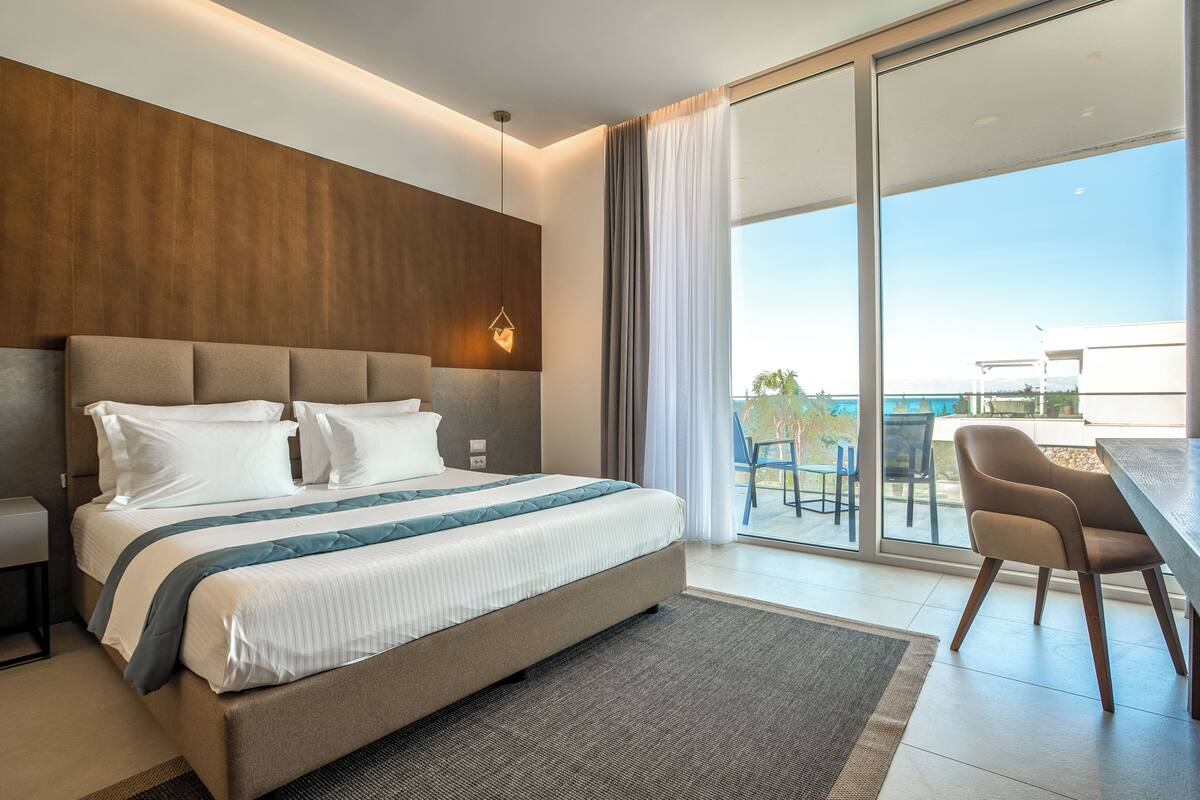 Una habitación de hotel moderna con una cama grande, un balcón con vista al mar y una zona de comedor con mesa y sillas.
