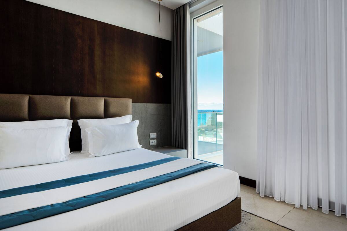 Una habitación de hotel moderna con una cama grande, cabecera de madera y vistas a la ciudad a través de amplias ventanas.