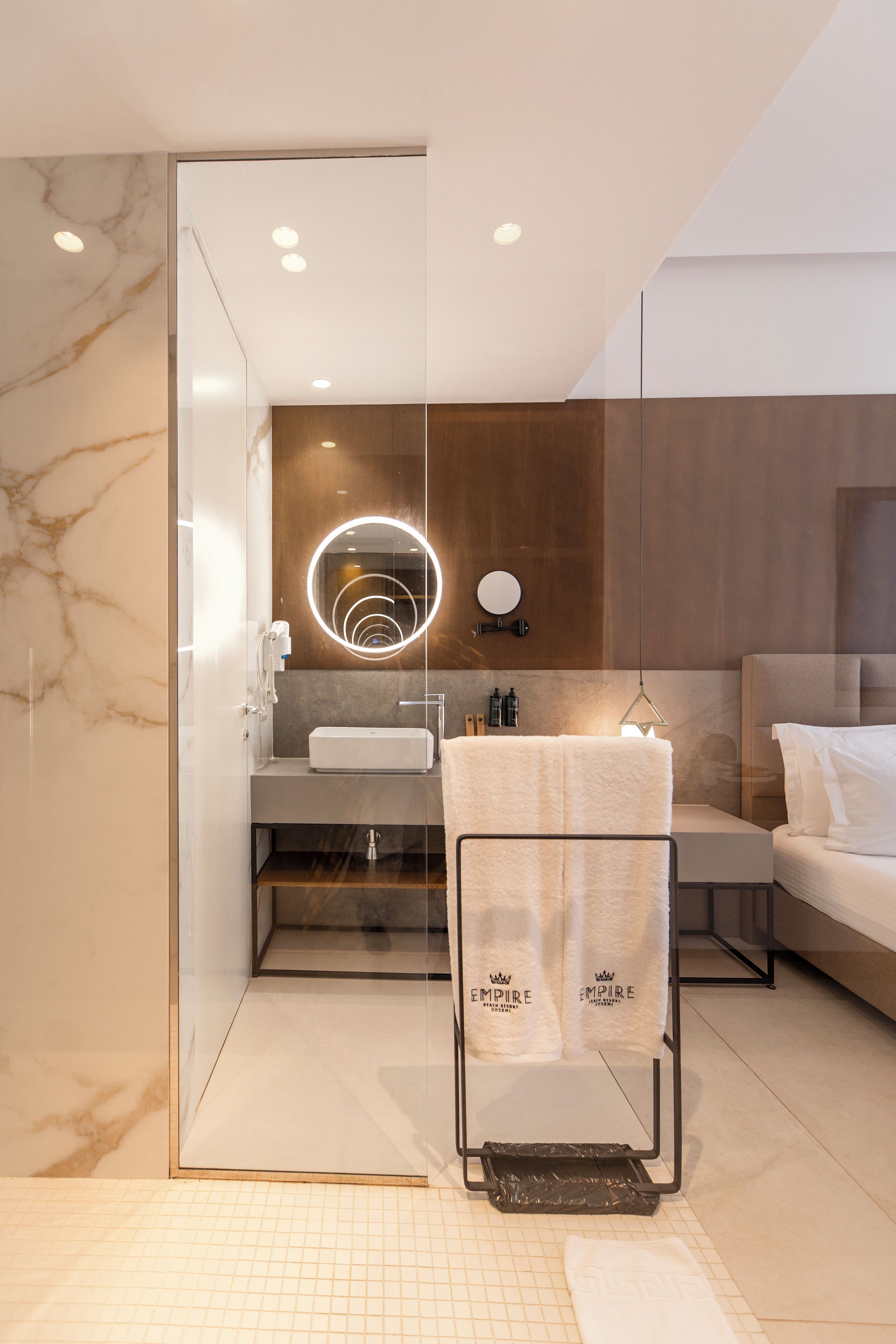 A modern bathroom with a marble wall, a glass shower enclosure, a round mirror with a light, a towel rack with towels, and a bed with white bedding.