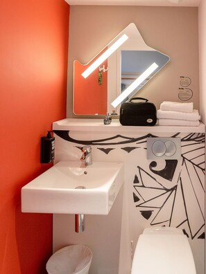 A modern bathroom with a white sink, a mirror with a guitar-shaped light, a black bag, and a toilet. - Ibis Budget Cognac (Chateaubernard)