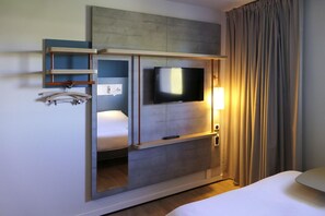 A hotel room with a bed, a TV mounted on a wall, a bedside table, a lamp, and a window with curtains. - Ibis Budget Cognac (Chateaubernard)
