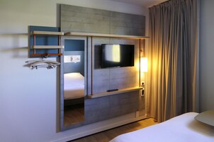 Quarto de hotel com cama, televisão na parede, mesa de cabeceira, abajur e janela com cortinas.