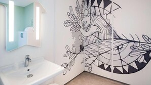 A bathroom with a white sink, a mirror, and a wall mural depicting a tree with leaves and branches. - Ibis Budget Cognac (Chateaubernard)