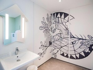 A bathroom with a white sink, a mirror, and a wall mural depicting a tree with leaves and branches. - Ibis Budget Cognac (Chateaubernard)