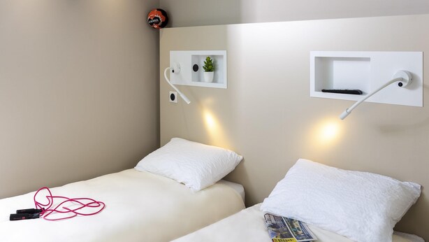 A modern hotel room with two single beds, a small shelf with a plant, a wall-mounted lamp, and a remote control. - Ibis Budget Cognac (Chateaubernard)