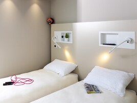 Quarto de hotel moderno com duas camas de solteiro, uma pequena prateleira com uma planta, uma luminária na parede e um controle remoto.