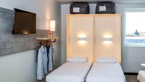 A modern hotel room with two single beds, a flat-screen TV mounted on the wall, a bedside table with a lamp, and a window with a view of the outside. - Ibis Budget Cognac (Chateaubernard)