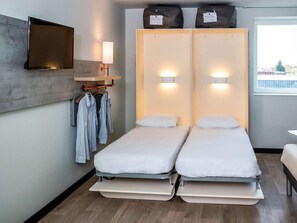 A modern hotel room with two single beds, a flat-screen TV mounted on the wall, a bedside table with a lamp, and a window with a view of the outside. - Ibis Budget Cognac (Chateaubernard)