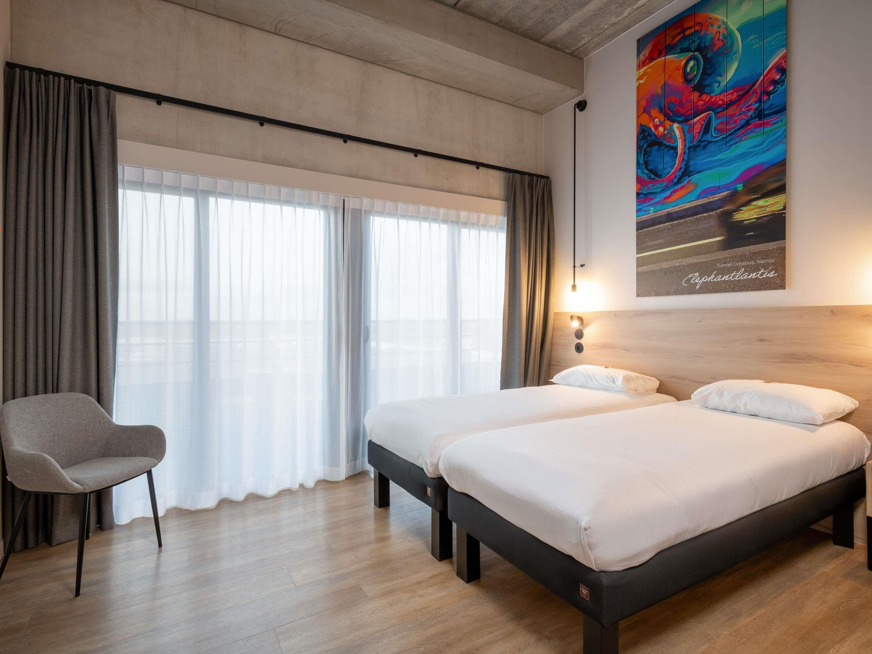 A hotel room with two beds, a chair, a large window with curtains, and a colorful painting on the wall.