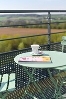 Una mesa con una taza de café y un libro en un balcón con barandilla de metal y vistas al campo.