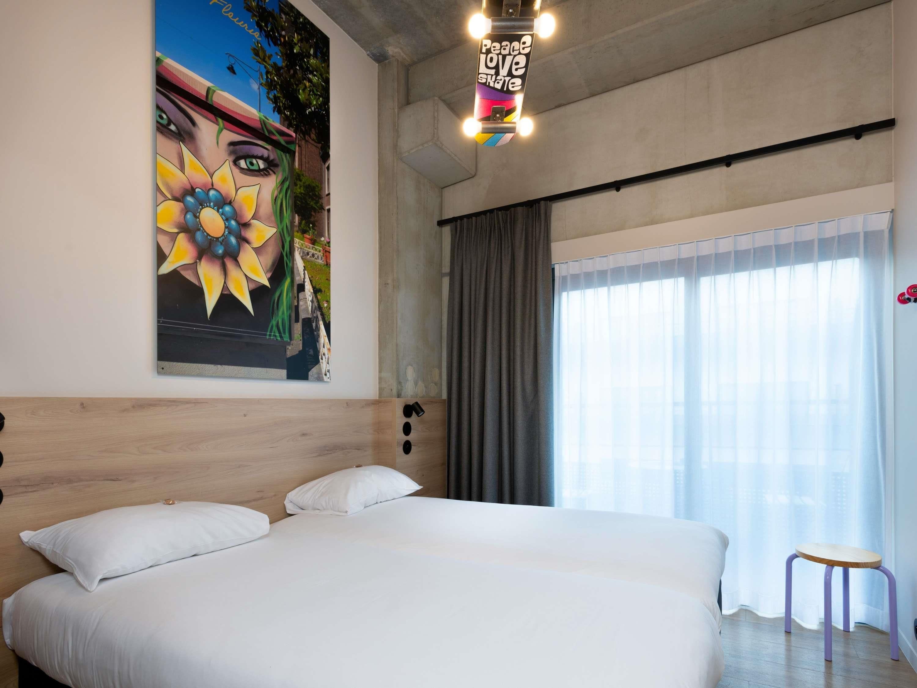 Una habitación de hotel moderna con una cama grande, un cuadro en la pared, una ventana con cortinas y un taburete pequeño.
