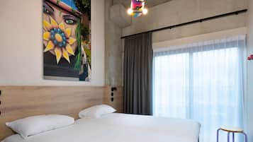 Una habitación de hotel moderna con una cama grande, un cuadro en la pared, una ventana con cortinas y un taburete pequeño.