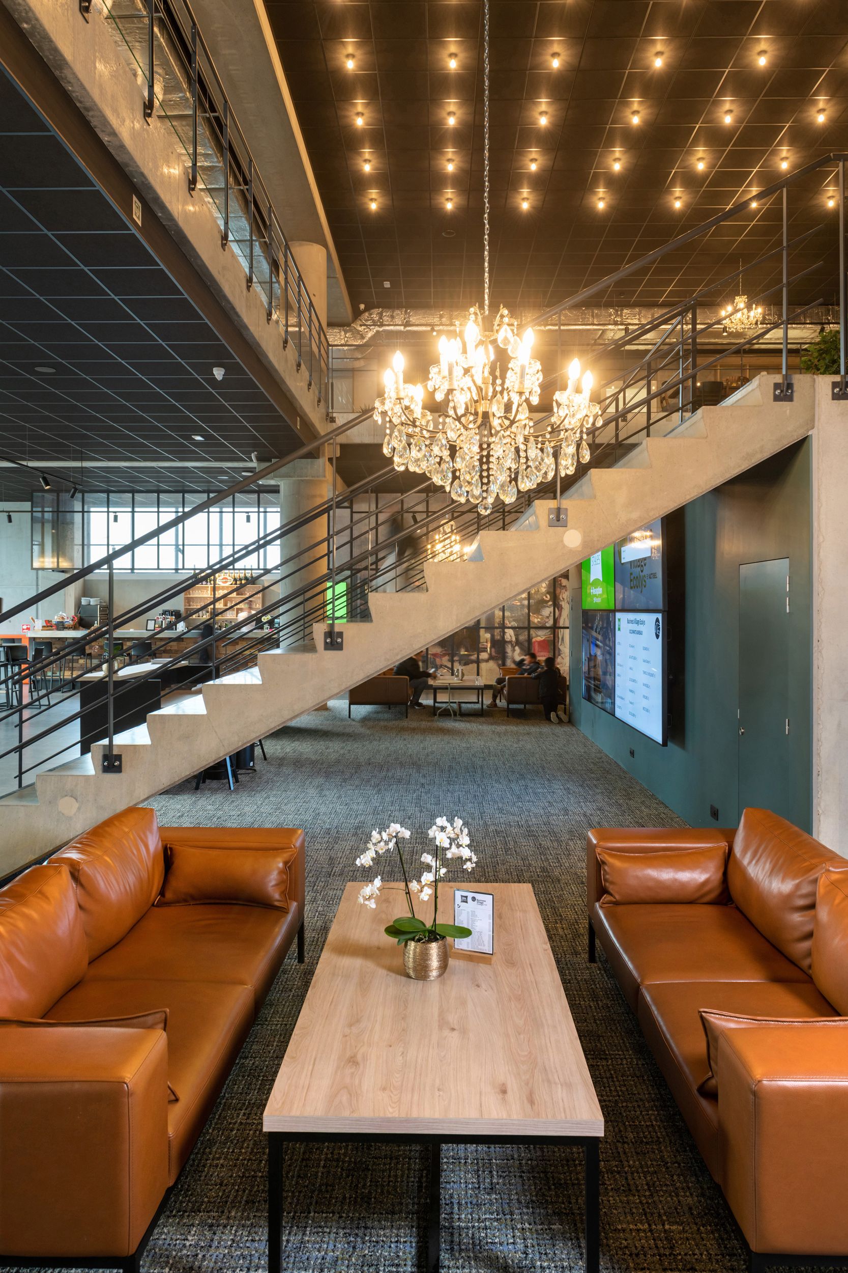 A spacious lobby with a chandelier, leather sofas, a wooden coffee table with orchids, and a staircase.