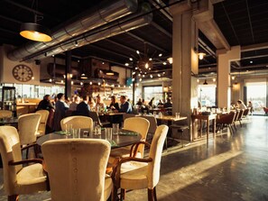 An indoor dining area with tables and chairs, a clock on the wall, and a view of the outside through large windows. - Ibis Styles Namur (Namur)