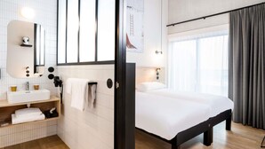 A hotel room with a bed, a bathroom with a sink and mirror, a wooden floor, and a window with curtains. - Ibis Styles Namur (Namur)