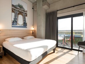 A hotel room with a large bed, a view of a cityscape, a balcony with chairs, and a painting of a bridge. - Ibis Styles Namur (Namur)