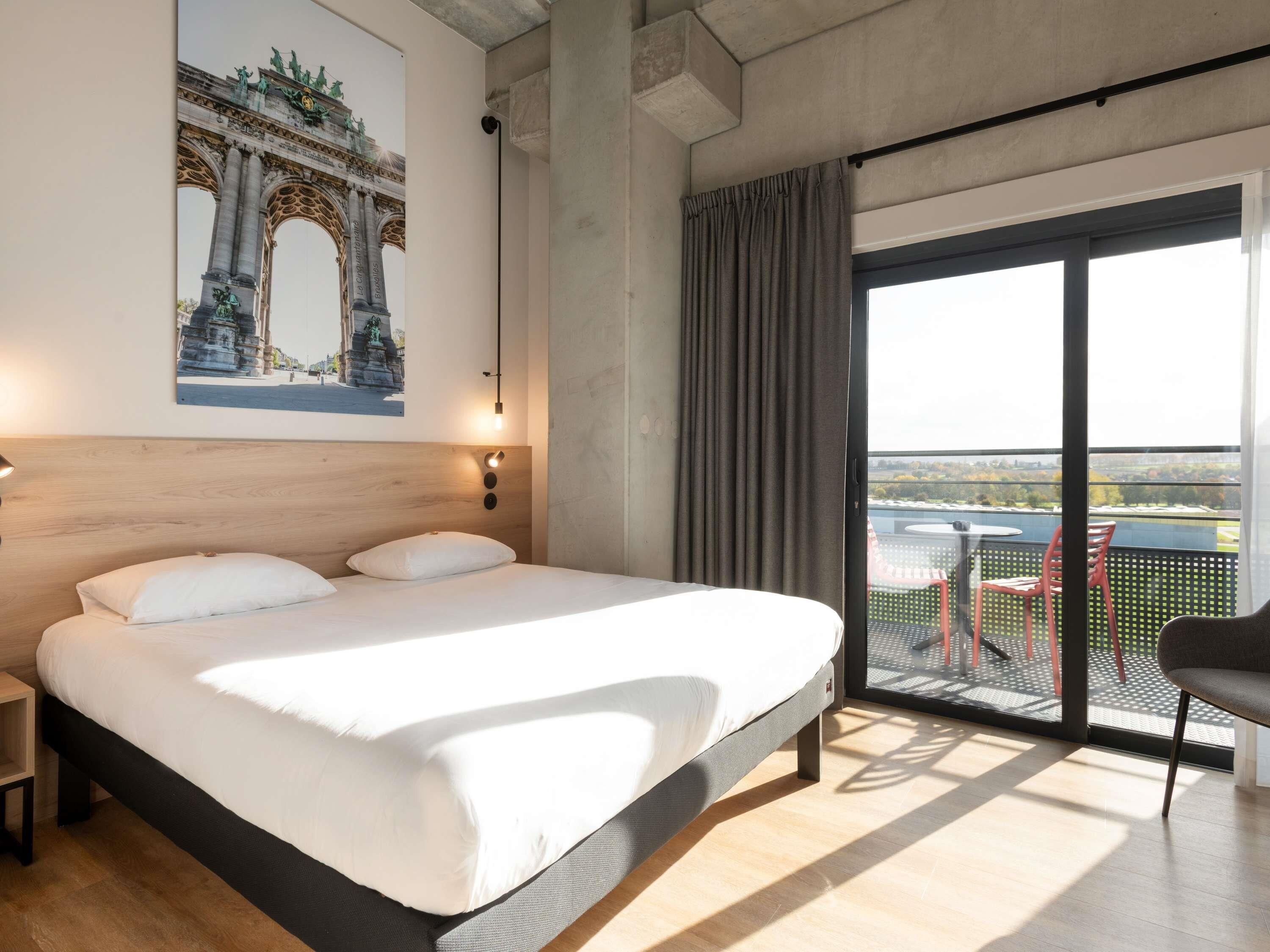 Hotelkamer met een groot bed, uitzicht op een stadsbeeld, een balkon met stoelen en een schilderij van een brug.