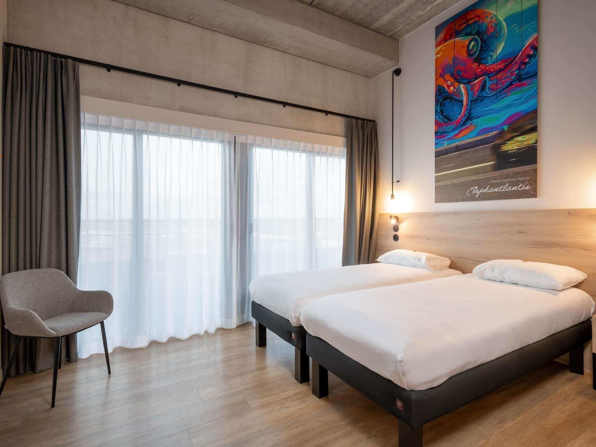 A hotel room with two beds, a chair, a large window with curtains, and a colorful painting on the wall.