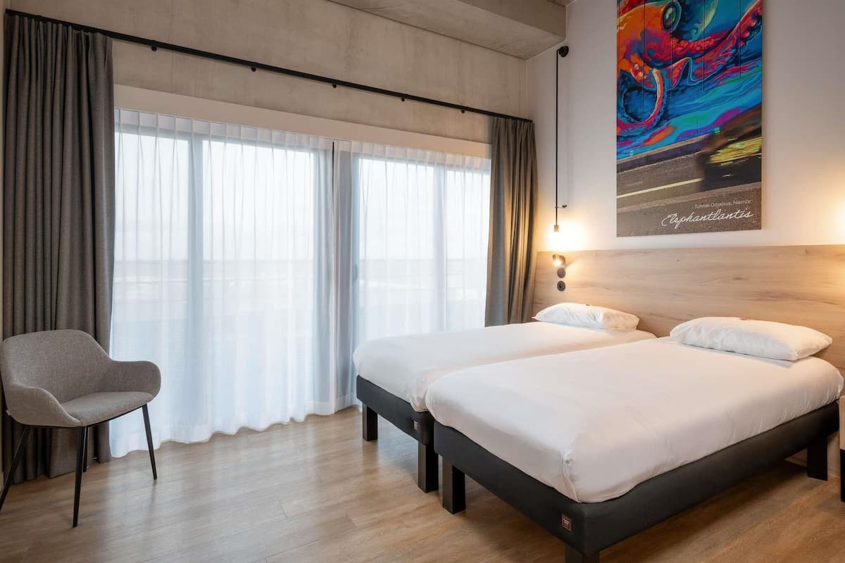 A hotel room with two beds, a chair, a large window with curtains, and a colorful painting on the wall.