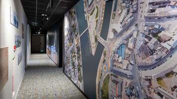Un couloir orné d’une grande fresque murale représentant un paysage urbain, doté d’un sol recouvert de moquette et de panneaux de signalisation.