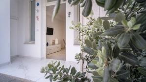 A hallway with a white door leading to a room with a bed, a dresser, and a TV. There are decorative items on the dresser and a plant in the foreground.