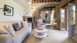 A cozy living room with a stone fireplace, a sofa with patterned cushions, a coffee table with magazines, and a chair with a striped cushion. - VILLA CANTUCCI (Lajatico)
