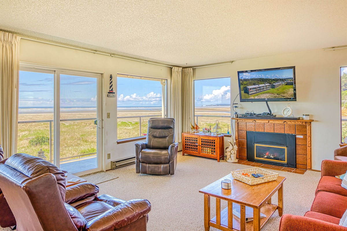 A spacious living room with a fireplace, a television, and a large window overlooking a beach.